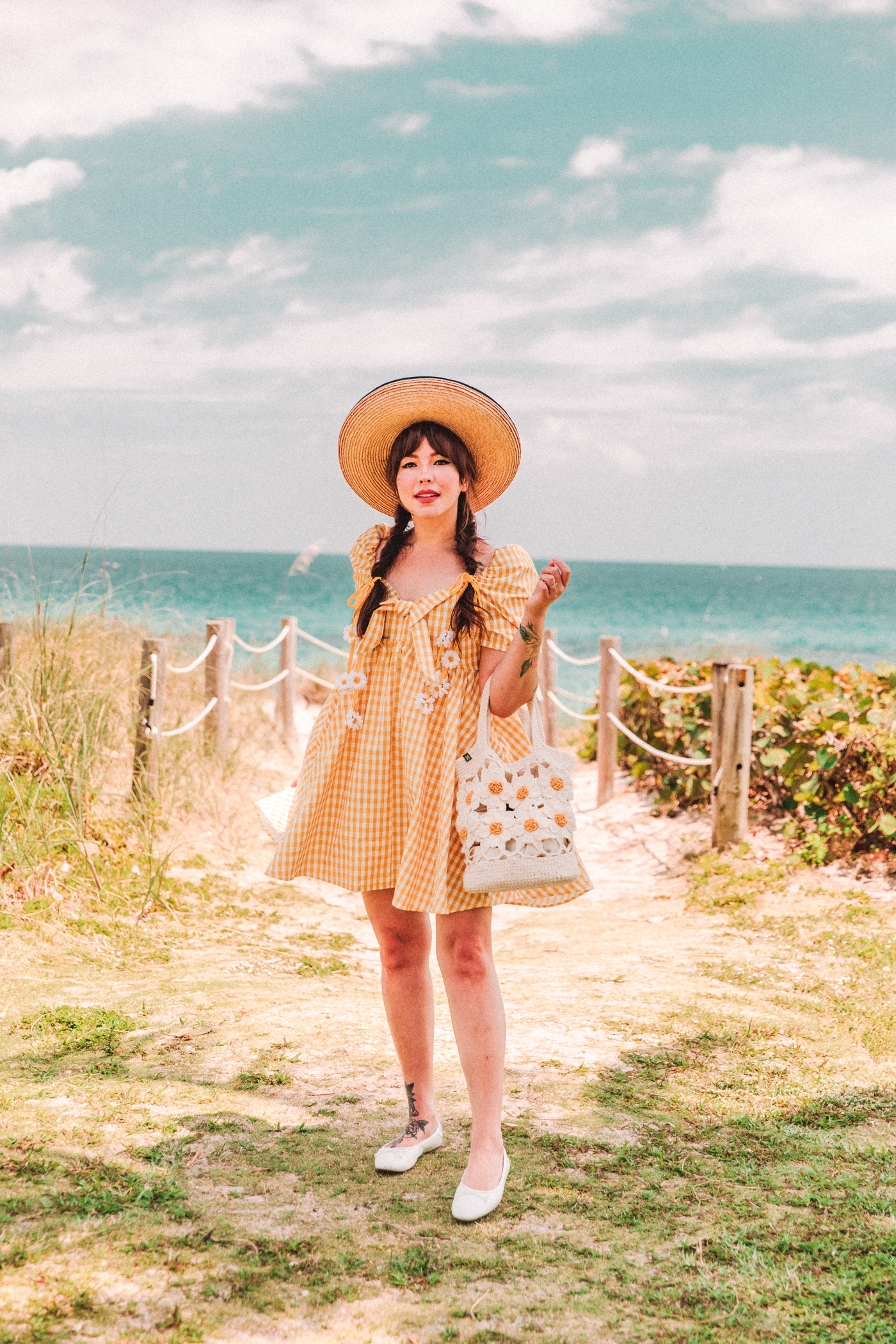 woman standing near the beach wearing yellow gingham dress, hat, and flats