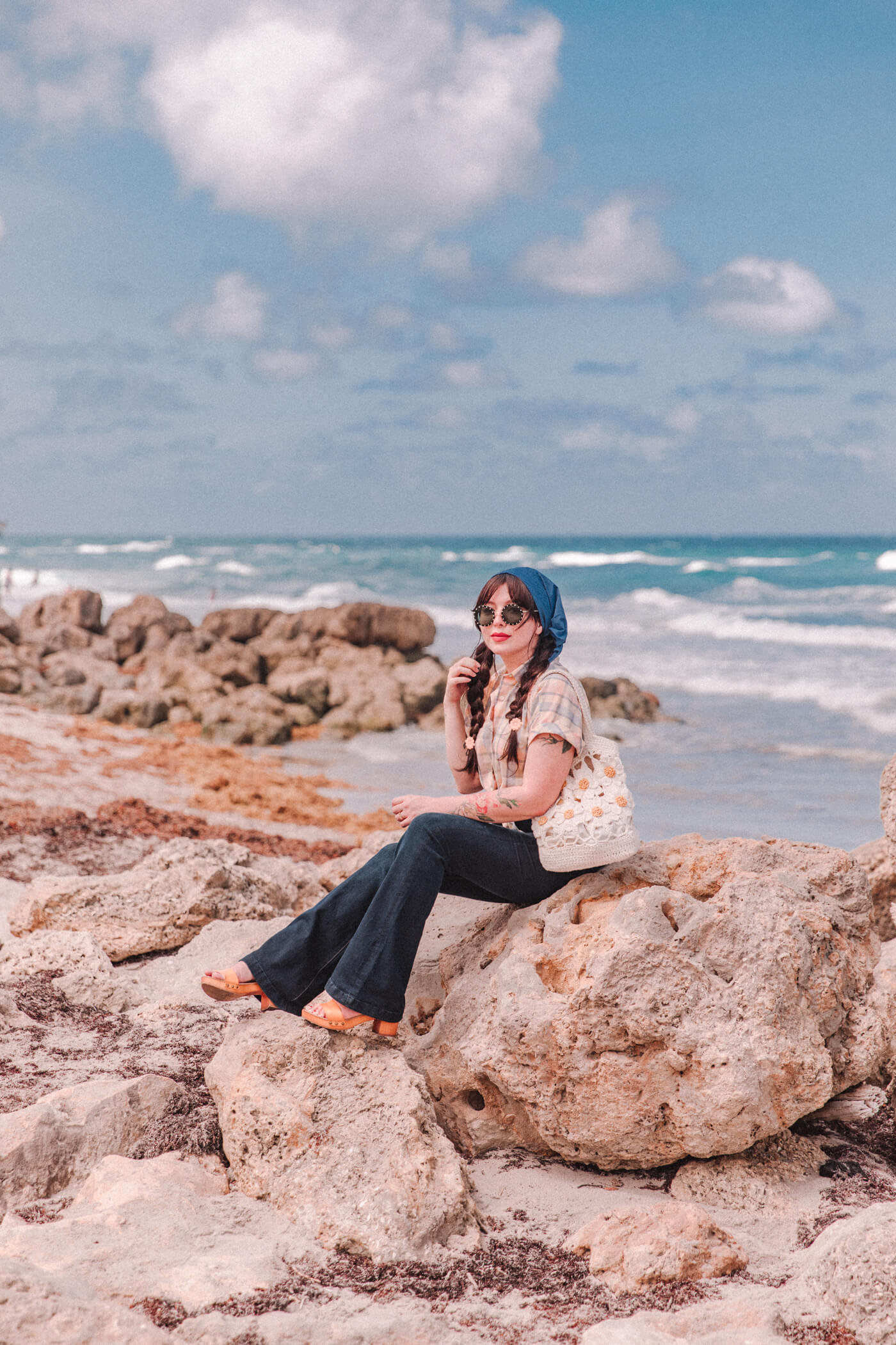 woman wearing bell bottom jeans and shirt and sitting on a rock near the beach