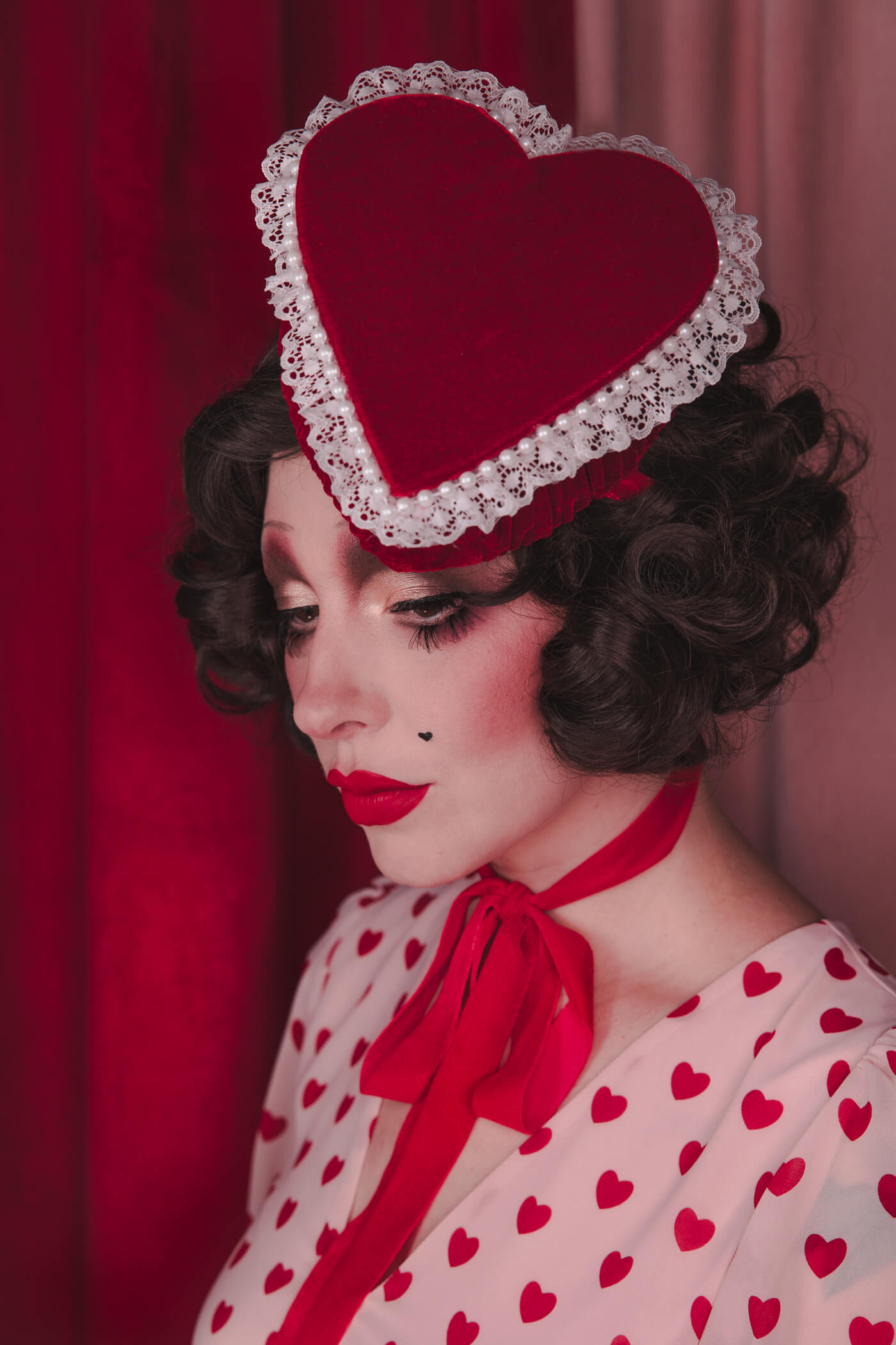 Pale girl with dark 20s 30s style hair and makeup, wearing a red velvet heart shaped box hat